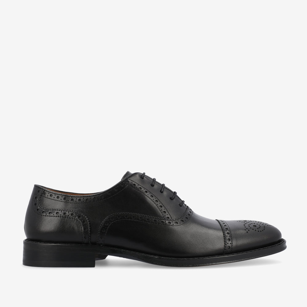 A black leather dress shoe with brogue detailing, featuring laces and a low heel, shown in a side profile against a white background.