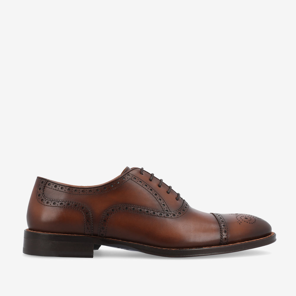 A brown leather Oxford dress shoe with brogue detailing and a low black heel, viewed from the side against a white background.