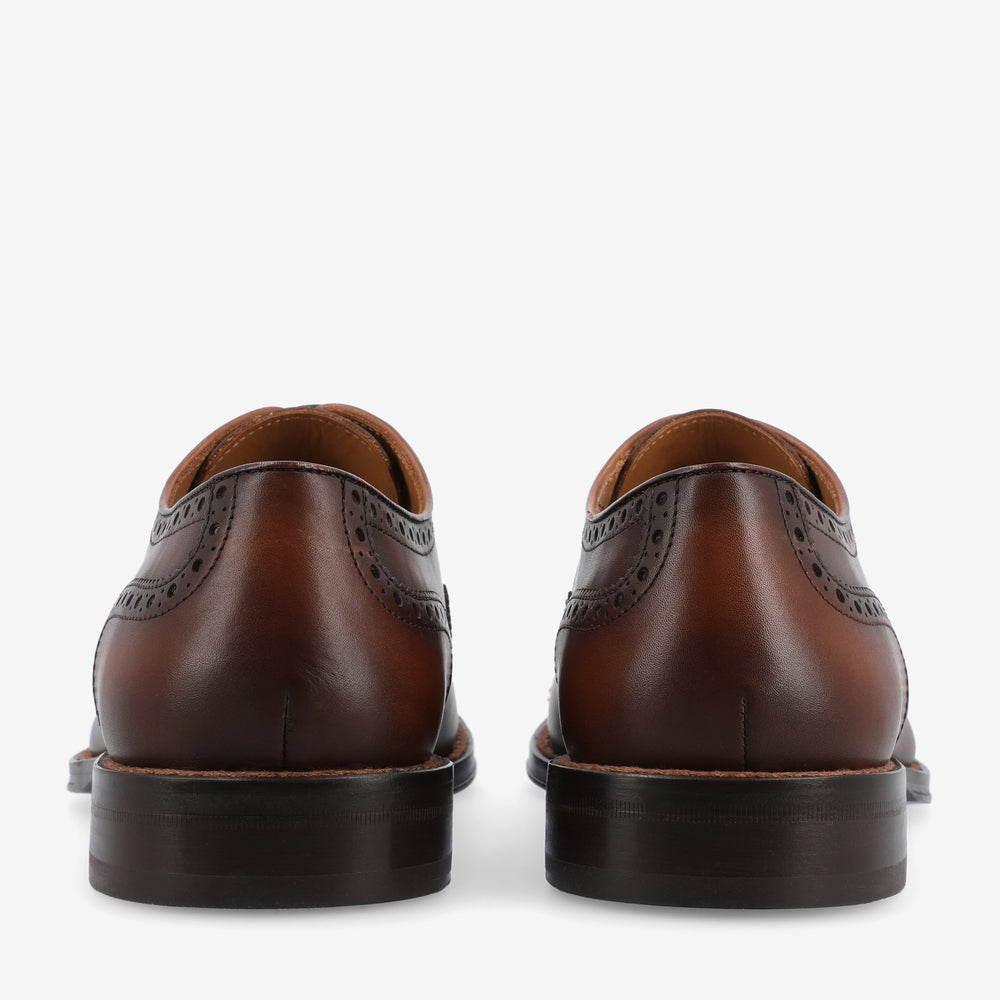 A pair of brown leather dress shoes with brogue detailing, viewed from the back.