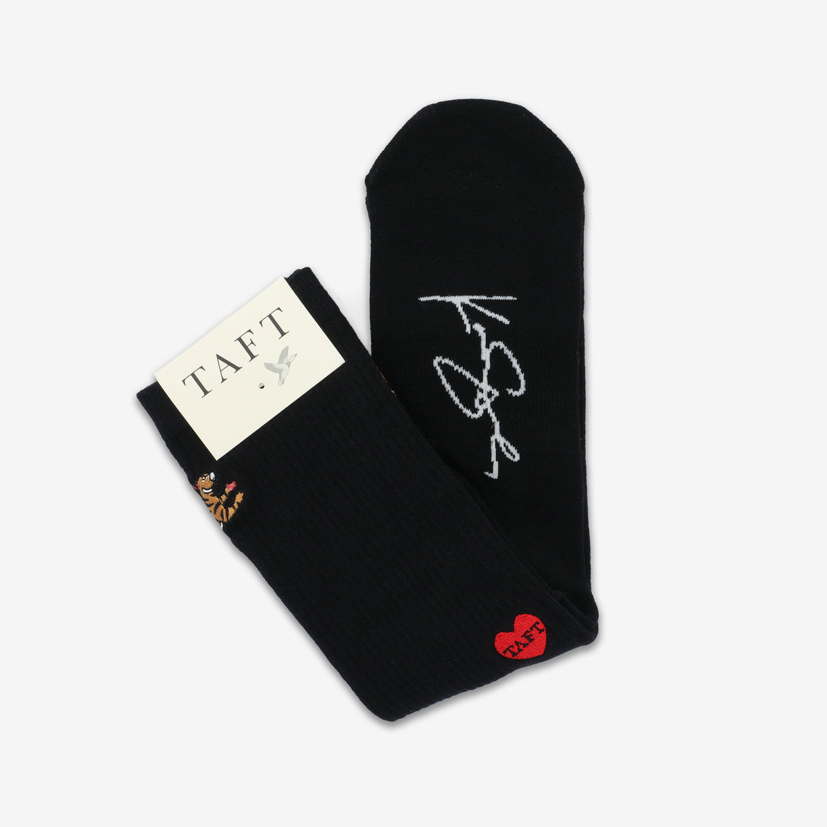 A pair of black socks with a white signature design and a red heart with TAFT lettering. Attached is a white tag labeled TAFT.