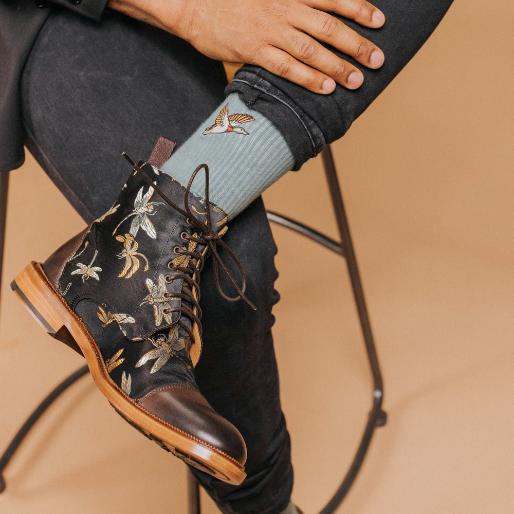 Close-up of a person sitting with one leg crossed, wearing black embroidered boots and light green socks featuring a butterfly design.