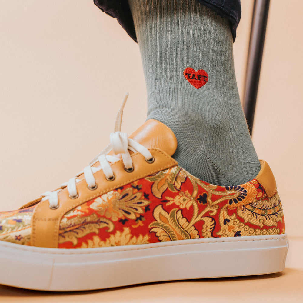 Close-up of a foot wearing a patterned sneaker with gold and orange floral designs and green socks featuring a small red heart with the word TAFT on it.