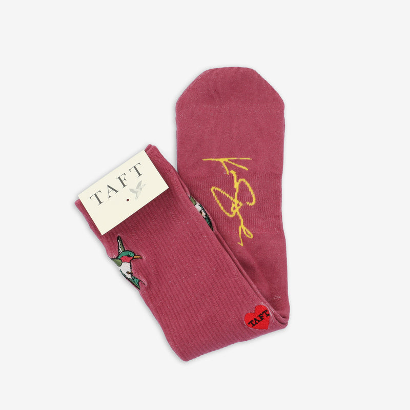 Pair of pink socks with embroidered birds, a yellow signature, and a small red heart label reading TAFT. A white cardboard tag also features the brand name TAFT.