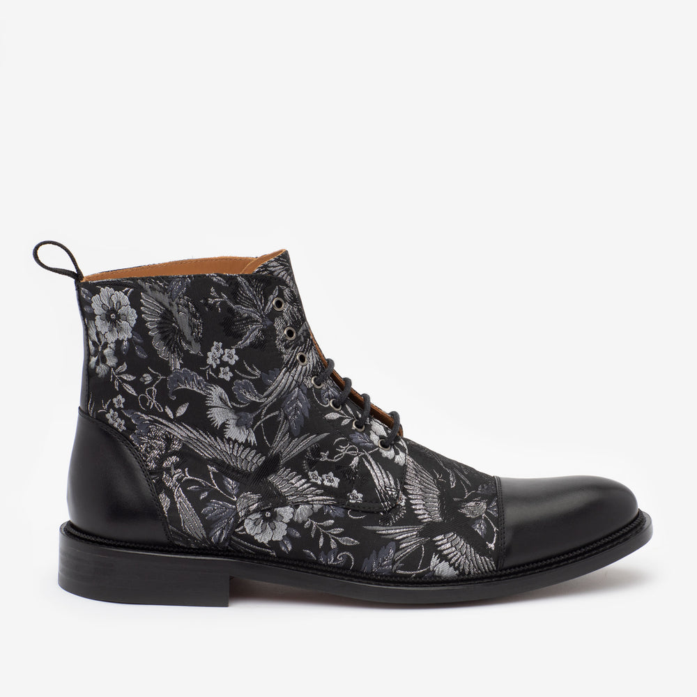 Black lace-up boot with a floral pattern, featuring a leather sole and a pull-tab on the heel. The main body showcases intricate floral designs in shades of gray.