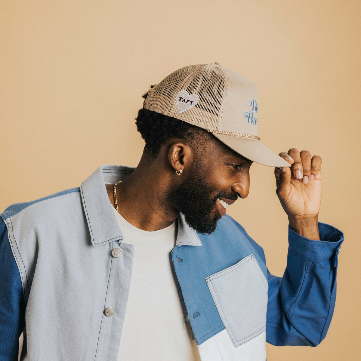 A man in a light-colored hat and blue jacket smiles while adjusting his hat. He stands against a beige background.