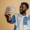 A man holds a beige cap with the phrase Deep Breaths embroidered in blue.