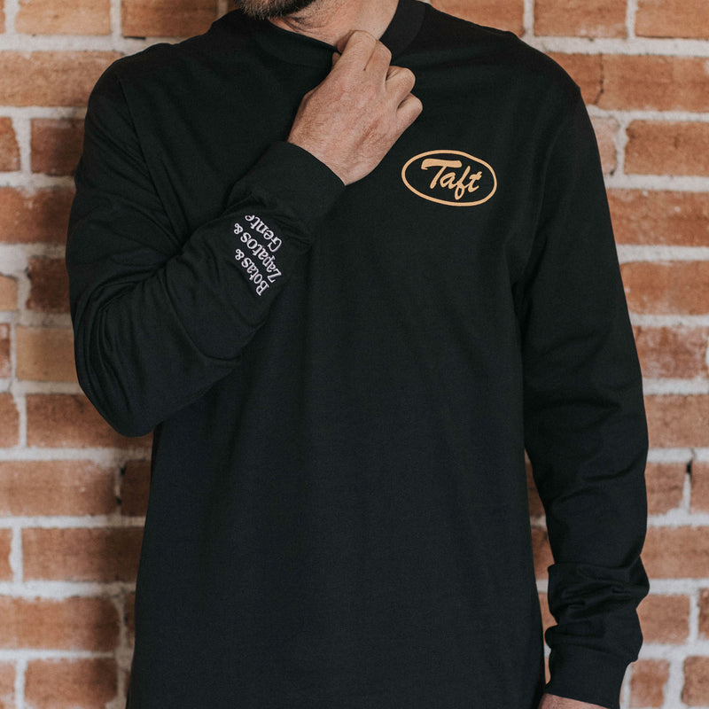 A person wearing a black long-sleeve shirt with the word Taft on the chest in yellow and text on the sleeve adjusts the collar, standing in front of a brick wall.