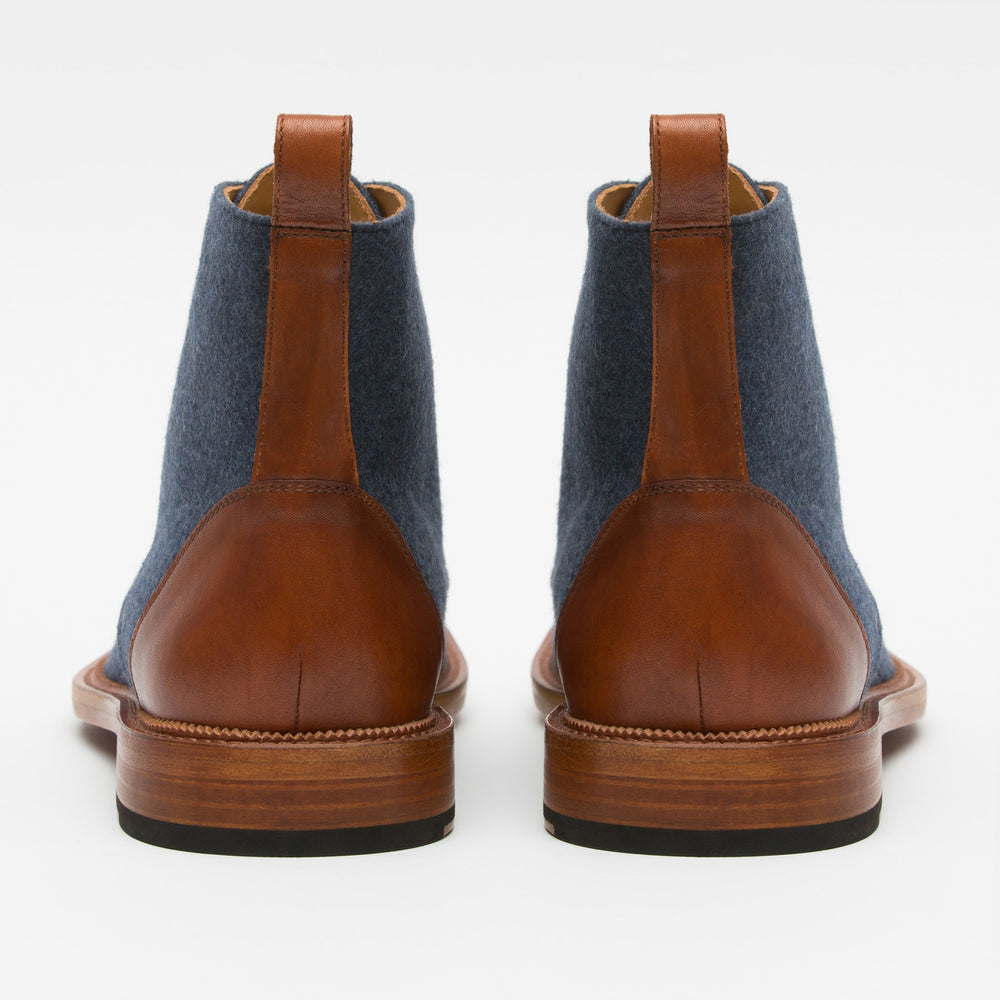 A pair of blue fabric boots with brown leather accents and pull tabs viewed from the back.