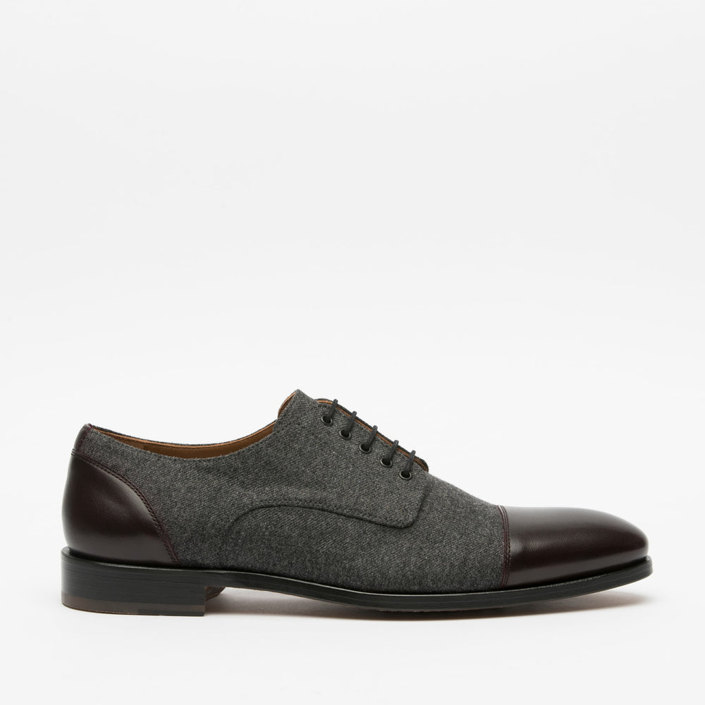 A grey and brown dress shoe with laces, featuring a leather toe and heel on a flat black sole, shown against a plain white background.