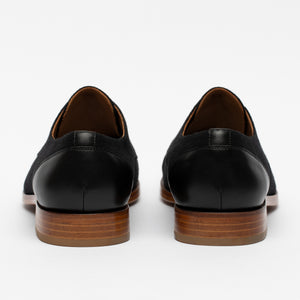 Rear view of a pair of black leather dress shoes with wooden soles on a white background.