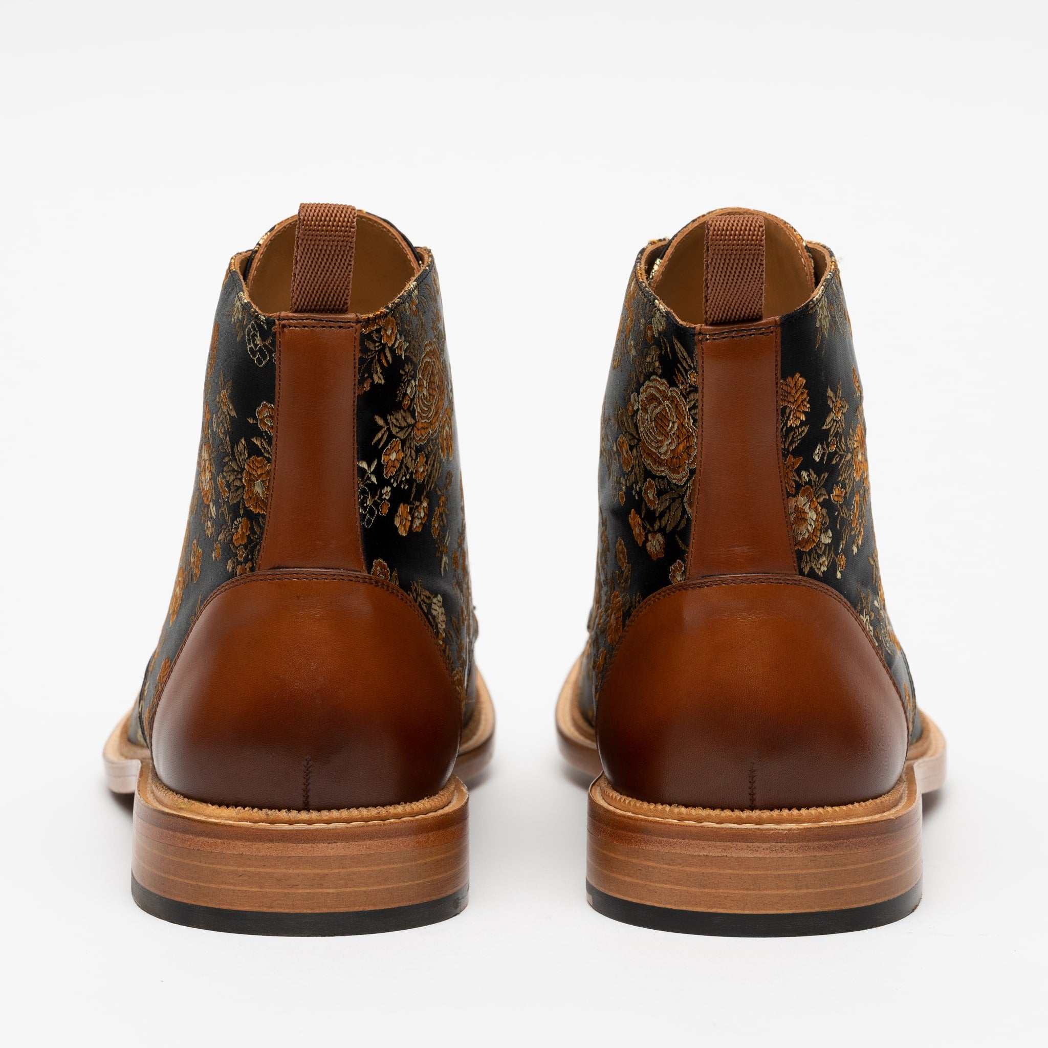 A pair of ankle-length boots viewed from the back, featuring brown leather and floral fabric patterns, with pull tabs at the top.