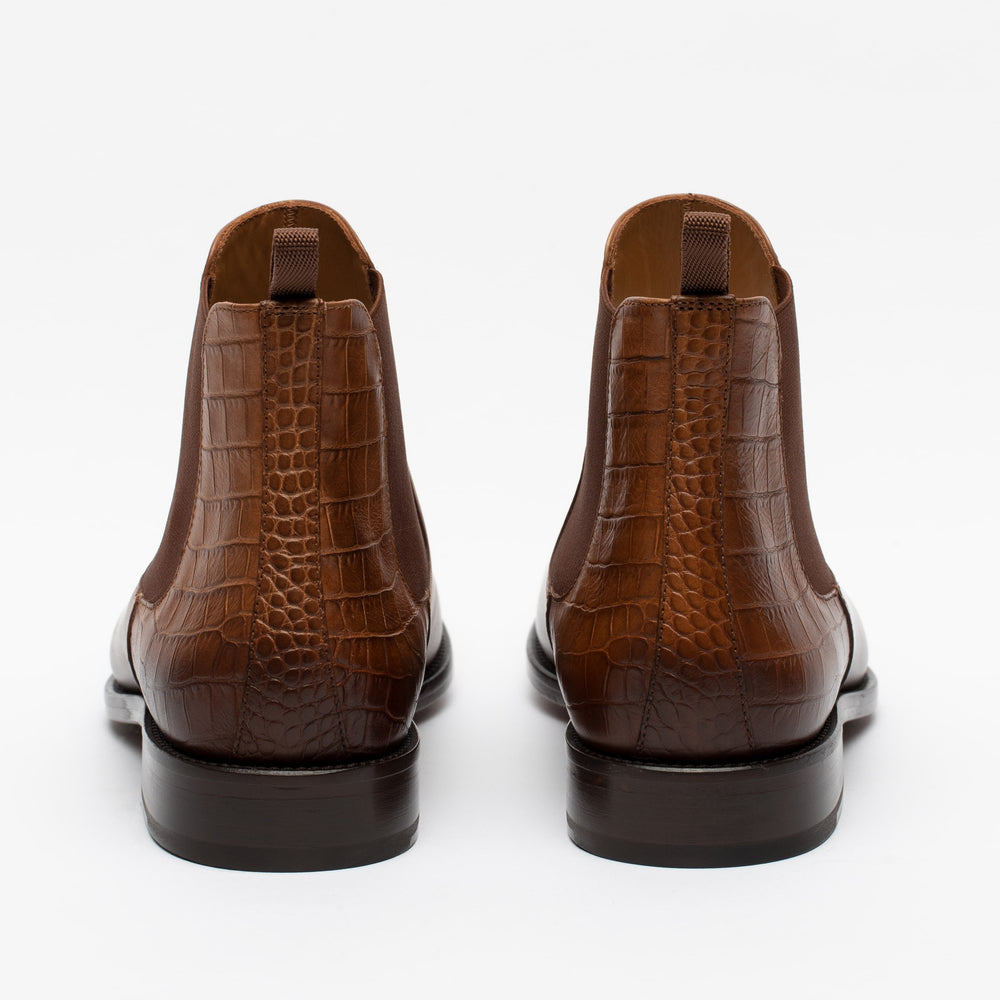 Rear view of brown, crocodile-patterned leather Chelsea boots with a pull-tab at the top of each boot, displayed against a white background.