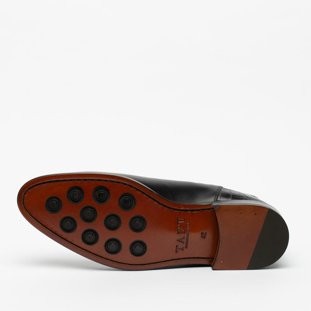 A single black dress shoe shown from the bottom, featuring a brown sole with multiple black rubber grip dots and a small logo that reads TAFT.