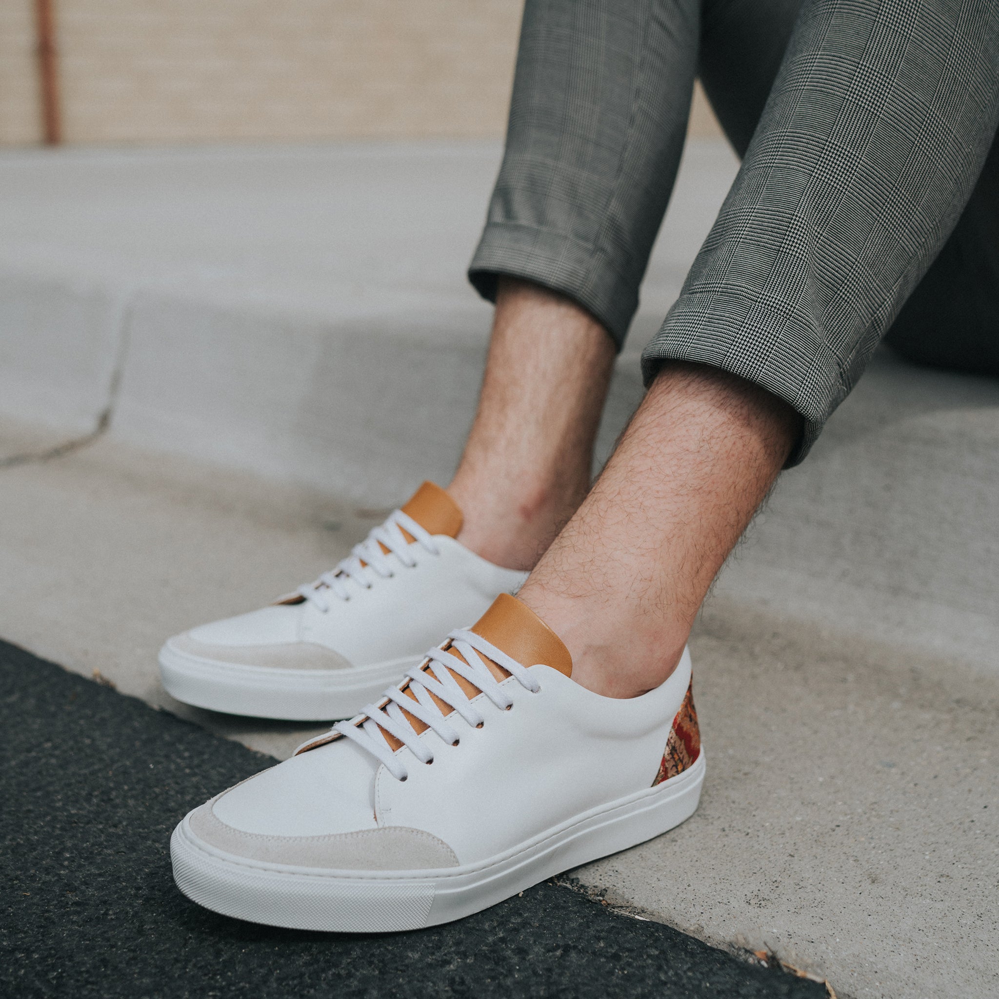 A person wearing white sneakers with brown accents, gray pants, and sitting on a curb.