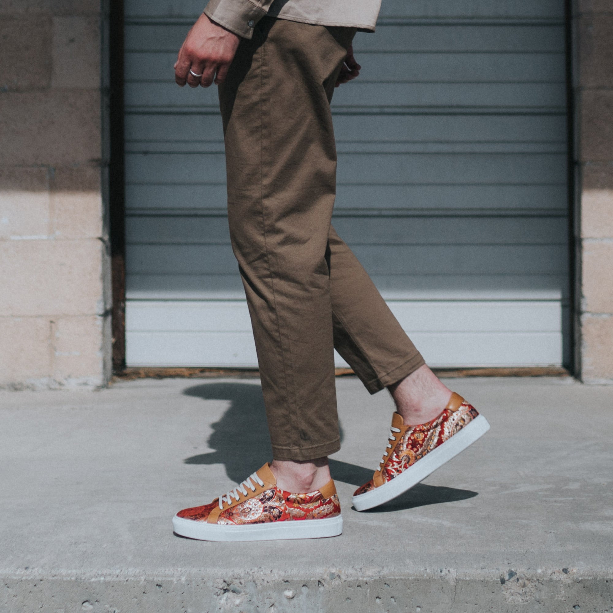 model walking in front of an old garage door wearing the sneaker in red paisley with brown pants