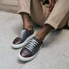 A person wearing beige pants and stylish sneakers with dark gray fabric and brown leather accents is seated on a concrete surface.