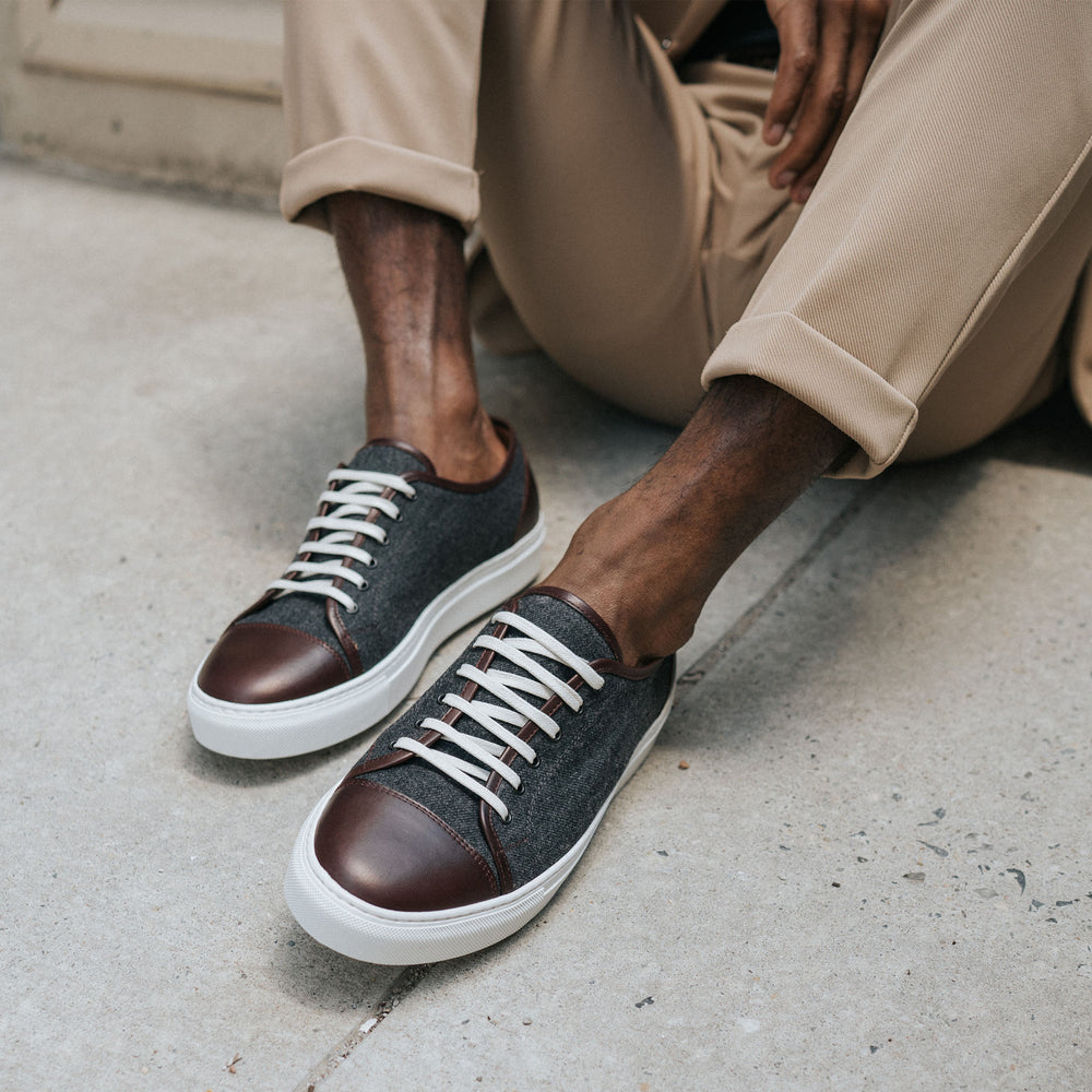 A person wearing beige pants and stylish sneakers with dark gray fabric and brown leather accents is seated on a concrete surface.