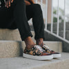 A person wearing black pants and floral-patterned sneakers sits on concrete steps with hands resting on knees.