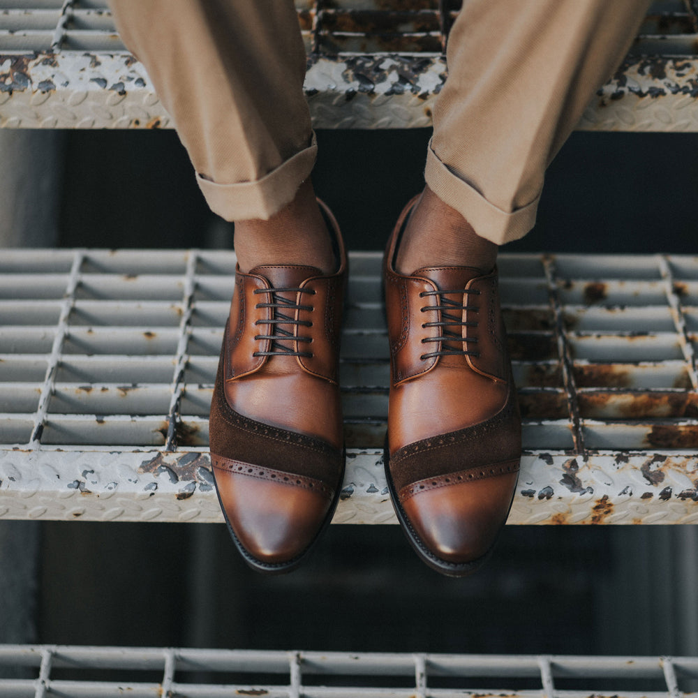 A person wearing brown leather Oxford shoes with dark brown accents, sitting on metal stairs with beige pants rolled up at the ankles.