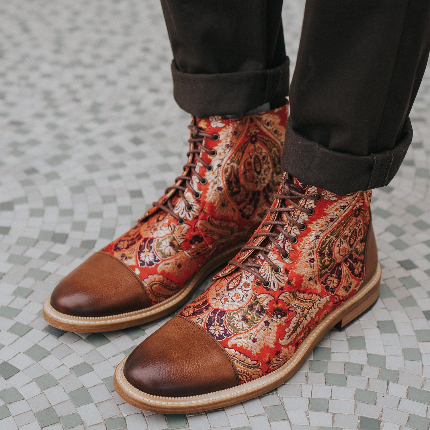 Person wearing patterned red and brown lace-up boots with brown leather caps and dark pants standing on a patterned tile floor.